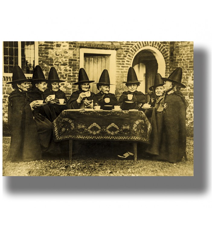 Vintage photograph of women dressed as witches sitting around a table and drinking tea outdoors.