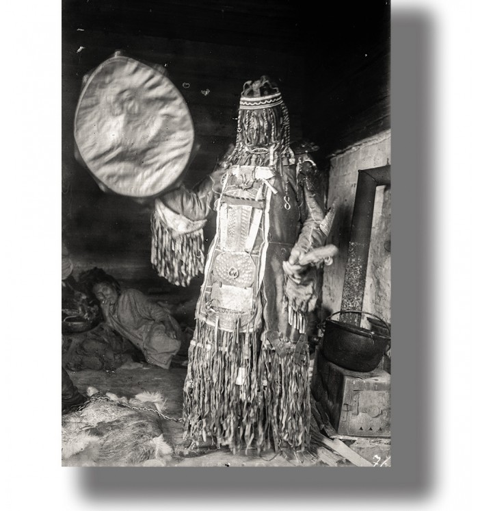 Vintage black and white photo of a Siberian shaman holding a drum inside a dwelling.
