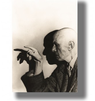 Bald man in profile pointing toward a wall, with a large dramatic shadow of his hand and face behind him.