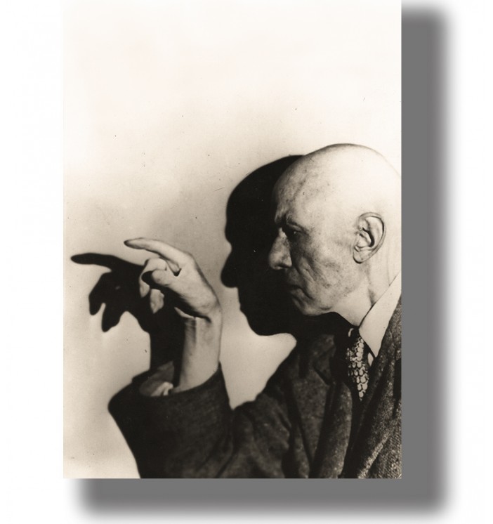 Bald man in profile pointing toward a wall, with a large dramatic shadow of his hand and face behind him.