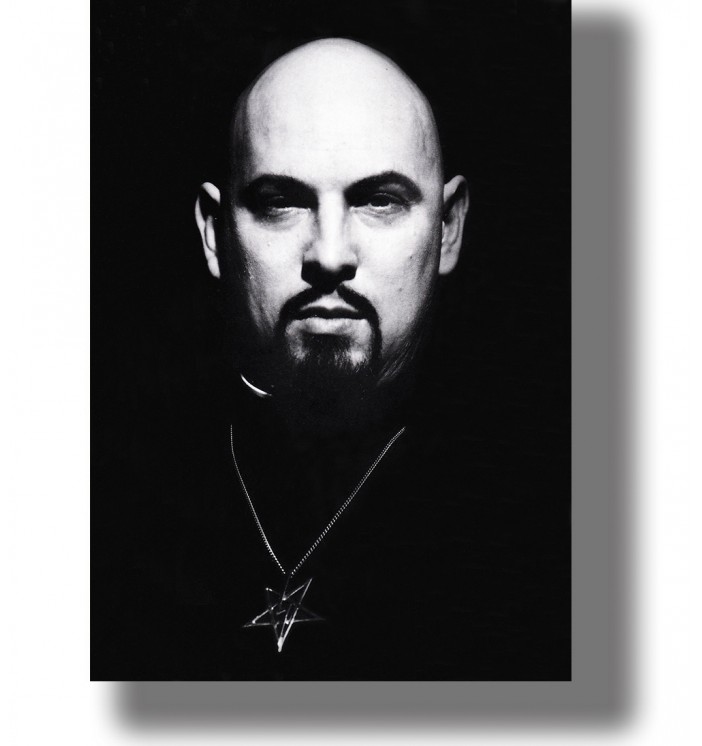 Black and white portrait of Anton LaVey with a shaved head, dark goatee and pentagram pendant against a black background.