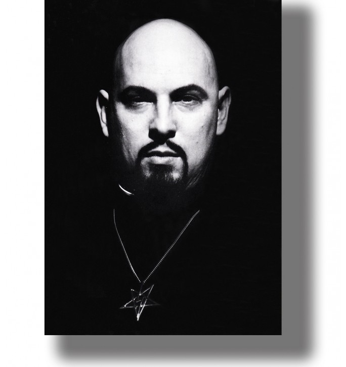 Black and white portrait of Anton LaVey with a shaved head, dark goatee and pentagram pendant against a black background.