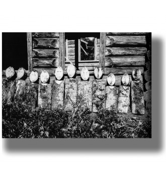 Row of wooden memorial figures standing in front of a rustic log house in a vintage black and white ethnographic photograph.