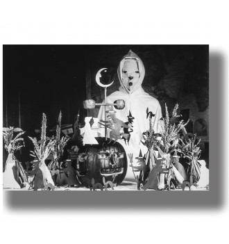 Masked figure in a white hooded Halloween costume standing behind a carved pumpkin and handmade witch decorations.