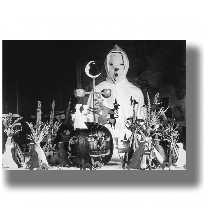 Masked figure in a white hooded Halloween costume standing behind a carved pumpkin and handmade witch decorations.