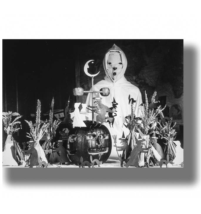 Masked figure in a white hooded Halloween costume standing behind a carved pumpkin and handmade witch decorations.