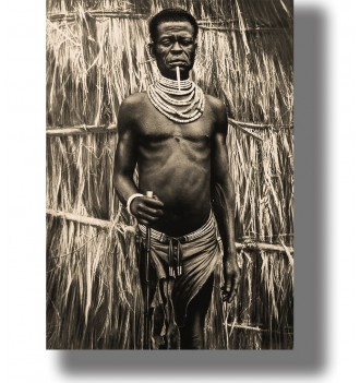 Vintage portrait of an African man standing before a thatched wall, wearing layered beaded necklaces and holding a staff.