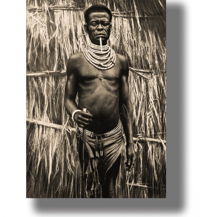 Vintage portrait of an African man standing before a thatched wall, wearing layered beaded necklaces and holding a staff.