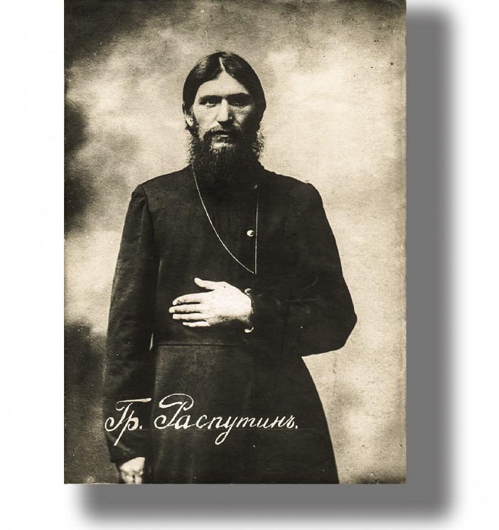 Old portrait of Grigori Rasputin standing in dark clothing with long beard and serious expression.