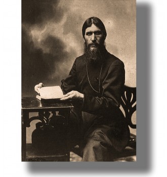 Vintage black and white portrait of Grigori Rasputin seated beside a table and holding a sheet of paper.
