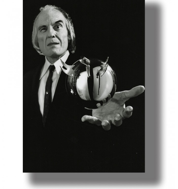 Black and white image of an older man in a dark suit holding a metallic flying sphere above his open hand.